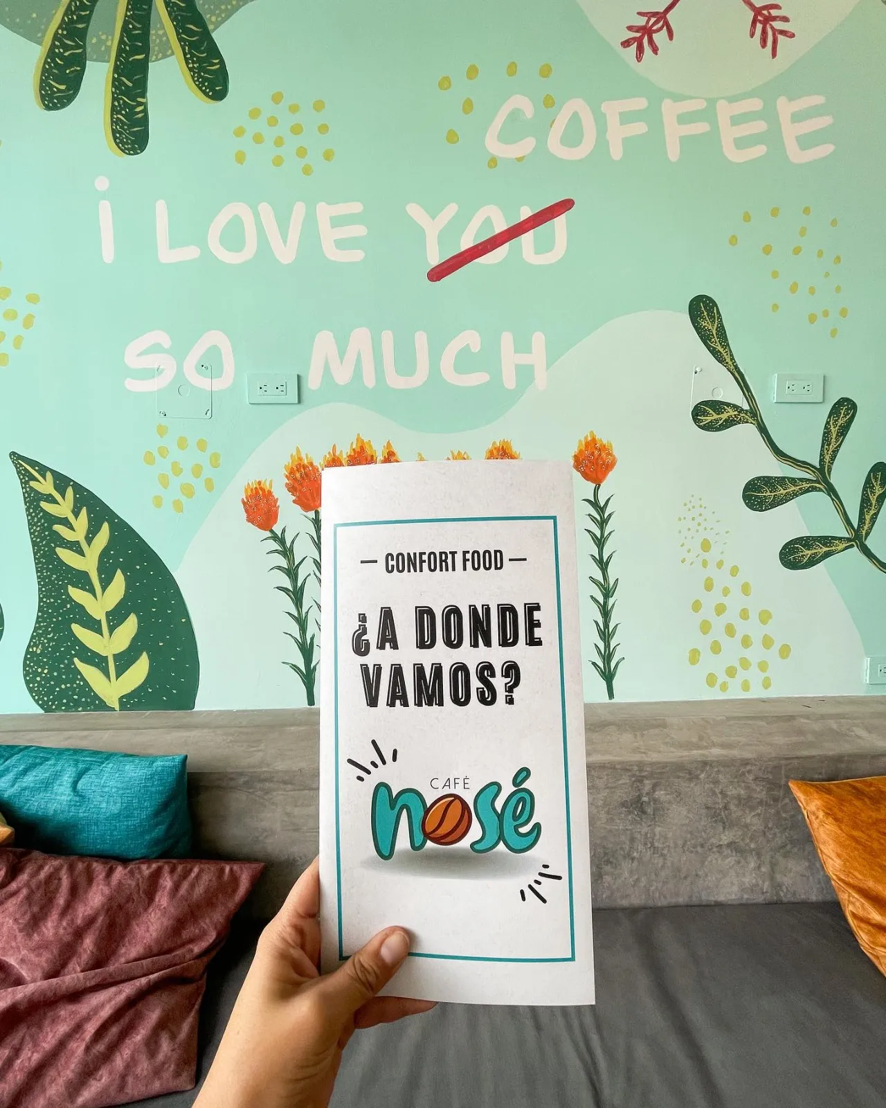 Una mano sostiene el menú de Café Nosé frente al mural turquesa del local