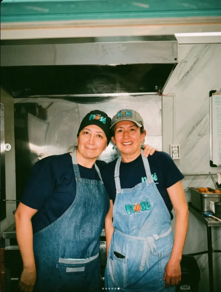 Dos compañeras de cocina abrazadas y sonriendo con gorras y delantales de Café Nosé